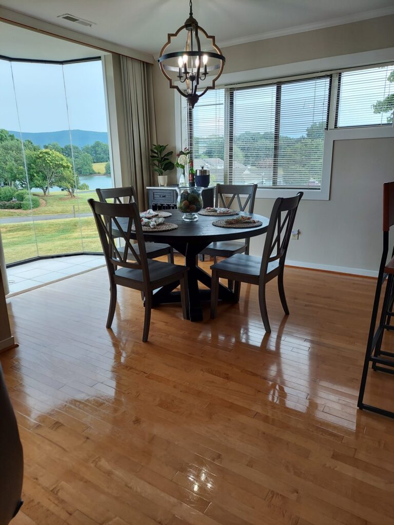 A dining area with a round table and four chairs by large windows overlooking a green lawn and trees.