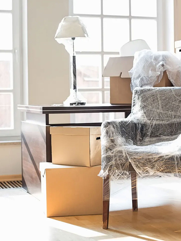 A desk, lamp, chair, and cardboard boxes wrapped in plastic, set in a bright room with large windows.