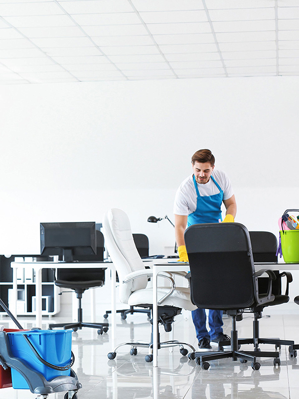 A person in an apron cleans an office, with desks, chairs, and cleaning supplies visible in the room.